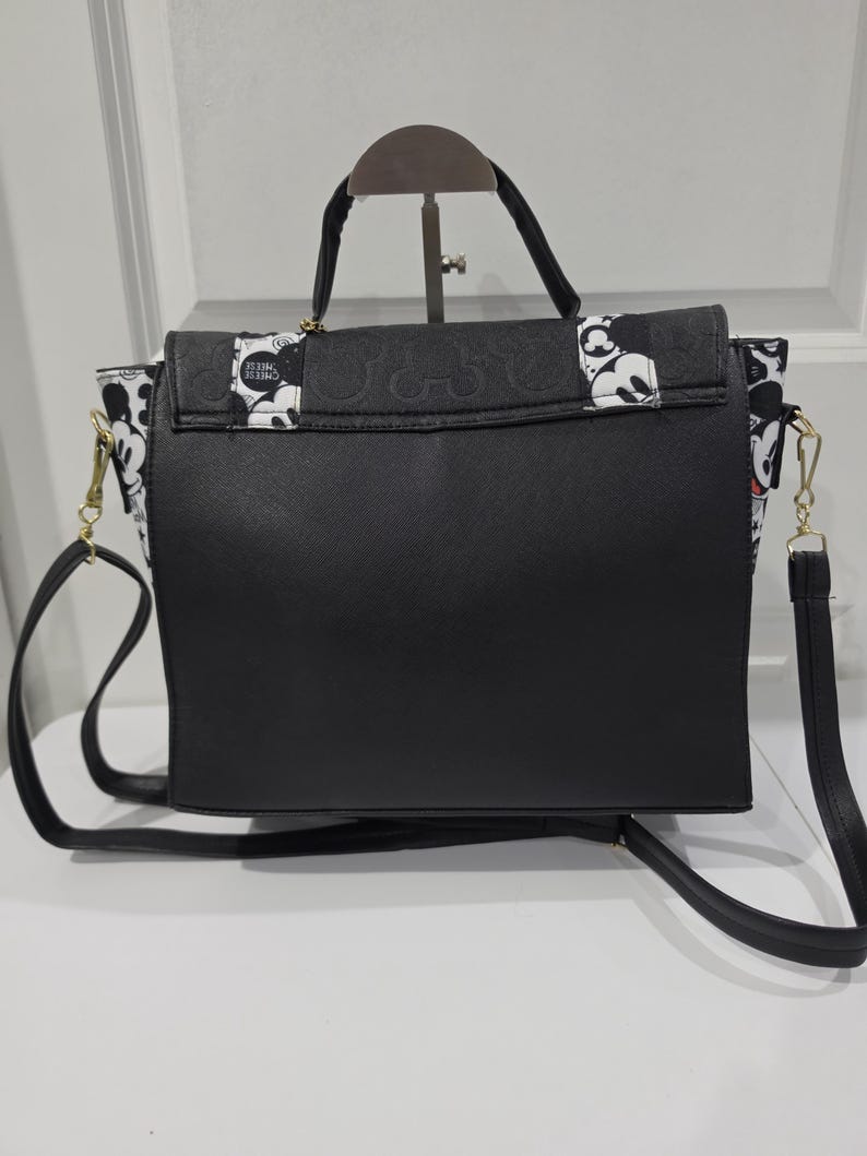 May include: A black handbag featuring a top handle and a detachable shoulder strap. The flap and side panels are decorated with a black and white pattern of Mickey Mouse faces and the text CHEESE. The main body of the bag is solid black.
