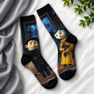 May include: A pair of black socks featuring characters from the animated film, "Coraline." One sock depicts a boy in a black coat, and the other shows a girl in a yellow coat holding flowers. The background includes a nighttime scene.