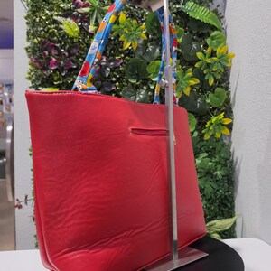 May include: A bright red tote bag featuring a horizontal zippered pocket on the exterior. The bag has two blue handles decorated with a repeating pattern of cartoon characters. The bag is suspended on a silver metal display stand.