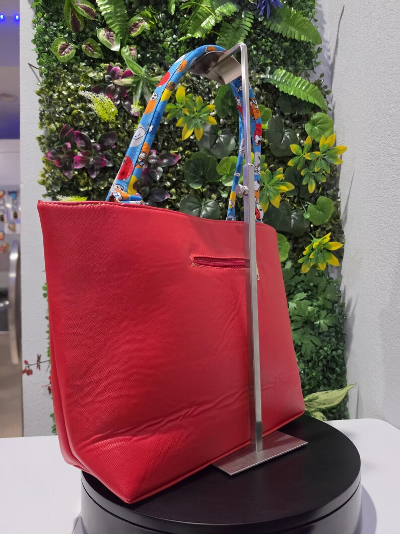 May include: A bright red tote bag featuring a horizontal zippered pocket on the exterior. The bag has two blue handles decorated with a repeating pattern of cartoon characters. The bag is suspended on a silver metal display stand.