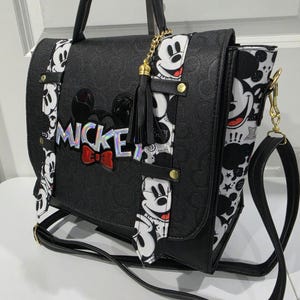 May include: A black satchel bag featuring a debossed Mickey Mouse pattern, a holographic MICKEY logo with a red bow, and two vertical white straps printed with Mickey Mouse faces. The bag includes a black tassel charm and a detachable shoulder strap.