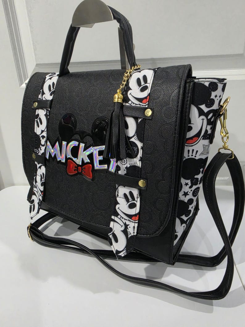 May include: A black satchel bag featuring a debossed Mickey Mouse pattern, a holographic MICKEY logo with a red bow, and two vertical white straps printed with Mickey Mouse faces. The bag includes a black tassel charm and a detachable shoulder strap.
