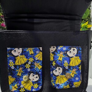 Puede incluir: Un bolso negro con dos bolsillos frontales con un estampado de personajes de dibujos animados en azul, amarillo y blanco. Los bolsillos tienen cierres magn&eacute;ticos dorados. El bolso tiene una solapa negra y un forro interior negro visible.