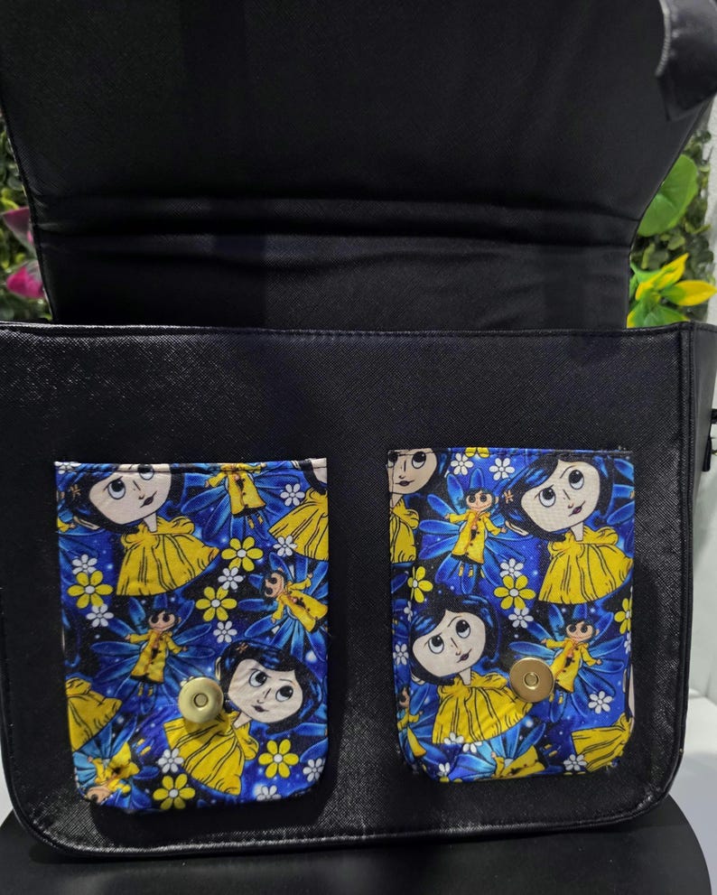 Puede incluir: Un bolso negro con dos bolsillos frontales con un estampado de personajes de dibujos animados en azul, amarillo y blanco. Los bolsillos tienen cierres magn&eacute;ticos dorados. El bolso tiene una solapa negra y un forro interior negro visible.