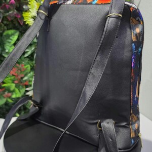 Puede incluir: Una mochila negra con acabado texturizado, correas finas para los hombros y herrajes met&aacute;licos en tono dorado. Los paneles laterales muestran un estampado gr&aacute;fico multicolor con tonos naranja, azul y morado, con cierre de cremallera.