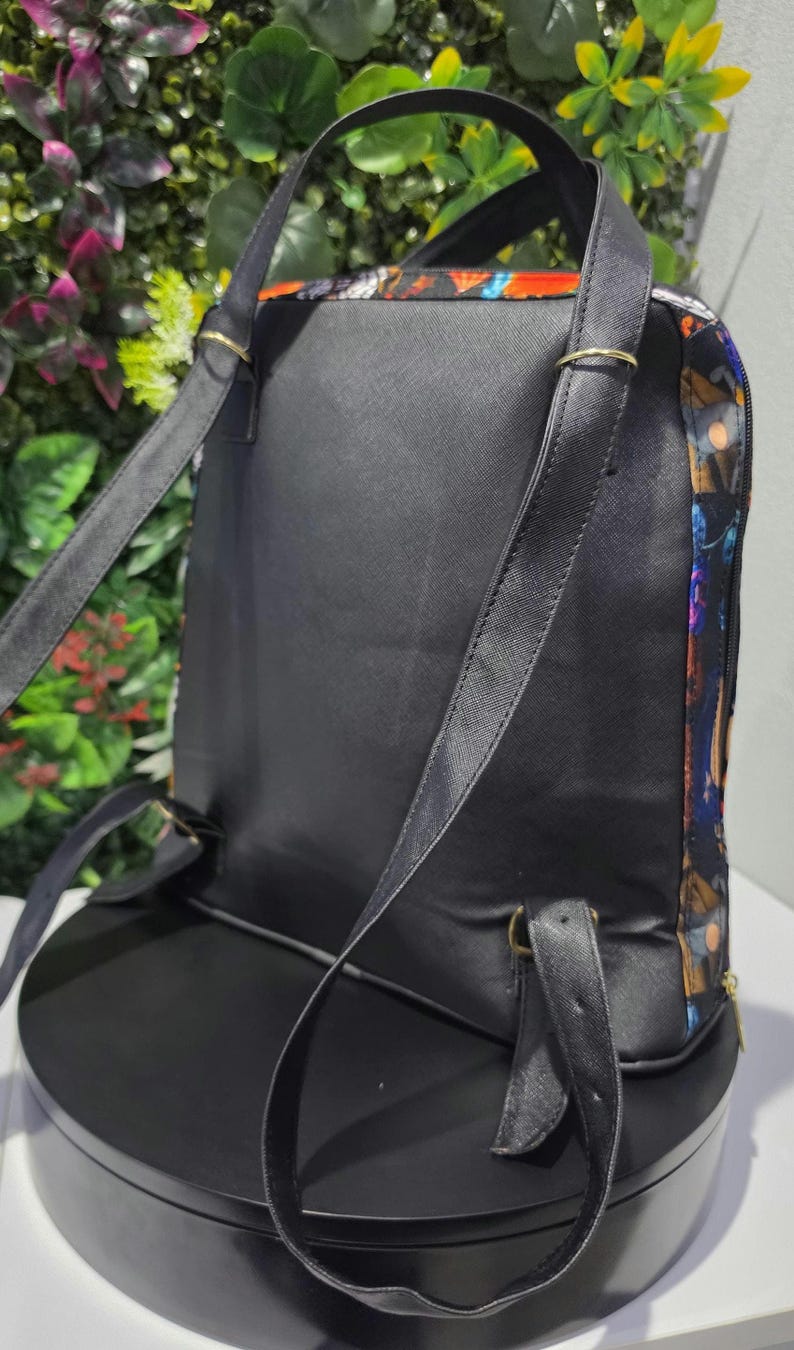 Puede incluir: Una mochila negra con acabado texturizado, correas finas para los hombros y herrajes met&aacute;licos en tono dorado. Los paneles laterales muestran un estampado gr&aacute;fico multicolor con tonos naranja, azul y morado, con cierre de cremallera.