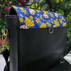 Puede incluir: Un bolso negro estructurado con asa superior y cierre de solapa. La solapa presenta una tela azul vibrante con un patr&oacute;n repetitivo de personajes de dibujos animados vestidos de amarillo y flores blancas. Incluye herrajes met&aacute;licos dorados.