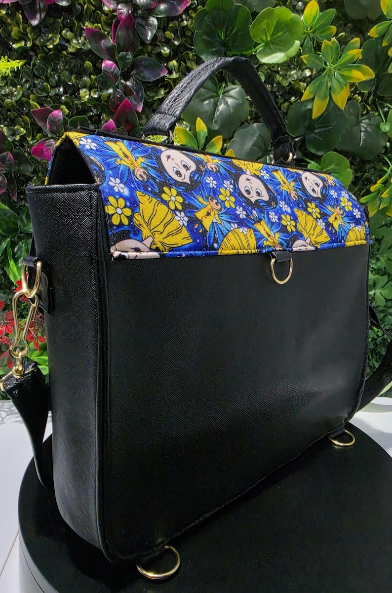 Puede incluir: Un bolso negro estructurado con asa superior y cierre de solapa. La solapa presenta una tela azul vibrante con un patr&oacute;n repetitivo de personajes de dibujos animados vestidos de amarillo y flores blancas. Incluye herrajes met&aacute;licos dorados.