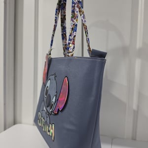 May include: A blue tote bag featuring a large, iridescent applique of the character Stitch with the name Stitch printed below in iridescent lettering. The bag has two shoulder straps decorated with a colorful, repeating character pattern.