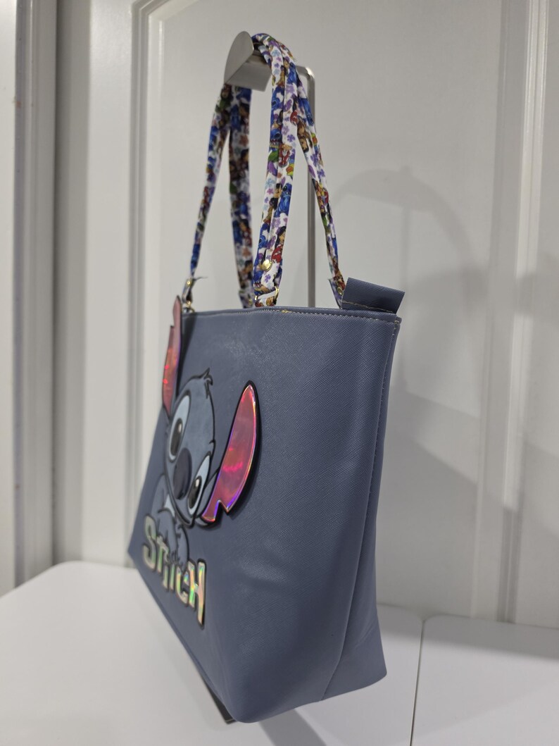 May include: A blue tote bag featuring a large, iridescent applique of the character Stitch with the name Stitch printed below in iridescent lettering. The bag has two shoulder straps decorated with a colorful, repeating character pattern.