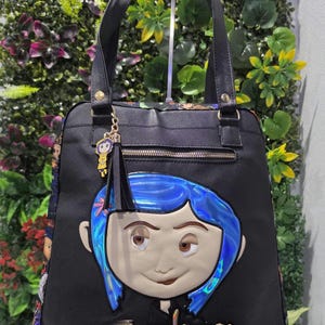 Puede incluir: Un bolso tote negro con un rostro de personaje grande, en relieve y con cabello azul iridiscente en la parte frontal. La palabra Coraline est&aacute; escrita en letras doradas debajo del rostro. Un dije y una borla negra cuelgan del asa.