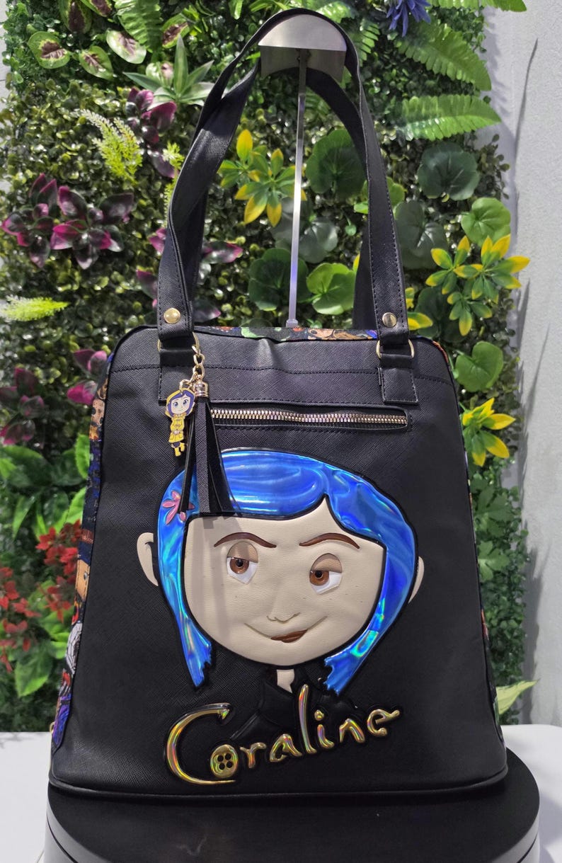 Puede incluir: Un bolso tote negro con un rostro de personaje grande, en relieve y con cabello azul iridiscente en la parte frontal. La palabra Coraline est&aacute; escrita en letras doradas debajo del rostro. Un dije y una borla negra cuelgan del asa.