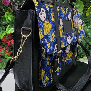 Puede incluir: Un bolso tipo cartera negro con un panel de tela azul y amarillo vibrante con ilustraciones de personajes de dibujos animados. El bolso incluye un asa superior, una correa de hombro desmontable con herrajes dorados y un dije con el texto Coraline.