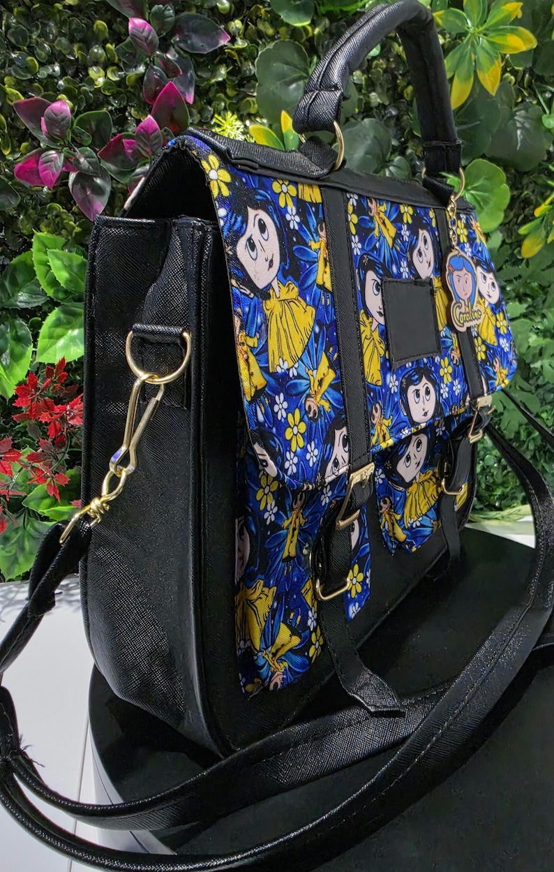 Puede incluir: Un bolso tipo cartera negro con un panel de tela azul y amarillo vibrante con ilustraciones de personajes de dibujos animados. El bolso incluye un asa superior, una correa de hombro desmontable con herrajes dorados y un dije con el texto Coraline.