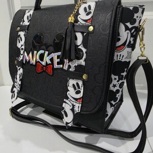 May include: A black satchel bag featuring a debossed Mickey Mouse pattern, a holographic MICKEY logo with a red bow, and white side panels with a black and white Mickey Mouse print. The bag includes a black tassel charm and a long shoulder strap.
