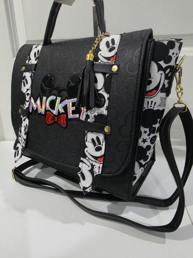 May include: A black satchel bag featuring a debossed Mickey Mouse pattern, a holographic MICKEY logo with a red bow, and white side panels with a black and white Mickey Mouse print. The bag includes a black tassel charm and a long shoulder strap.