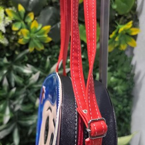May include: A round black crossbody bag featuring a character graphic on the front panel. The bag has a red zipper closure, red adjustable straps with white stitching, and silver-toned metal hardware, including a buckle and rivets on the strap.