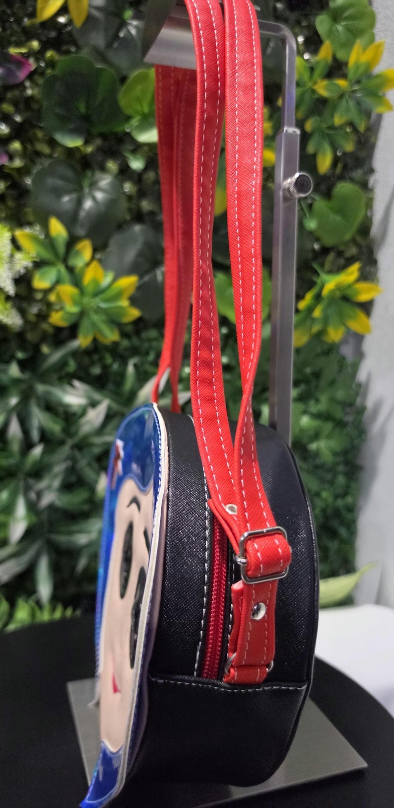 May include: A round black crossbody bag featuring a character graphic on the front panel. The bag has a red zipper closure, red adjustable straps with white stitching, and silver-toned metal hardware, including a buckle and rivets on the strap.