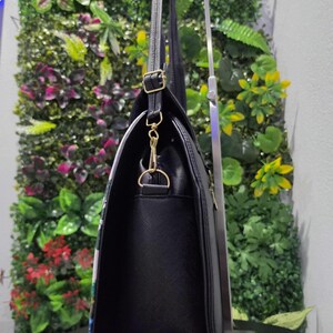 May include: A black handbag with a textured surface, featuring gold-toned metal hardware including a side D-ring, a swivel clasp, and an adjustable strap buckle. The bag is displayed in profile, showing its vertical orientation and structured side panel.