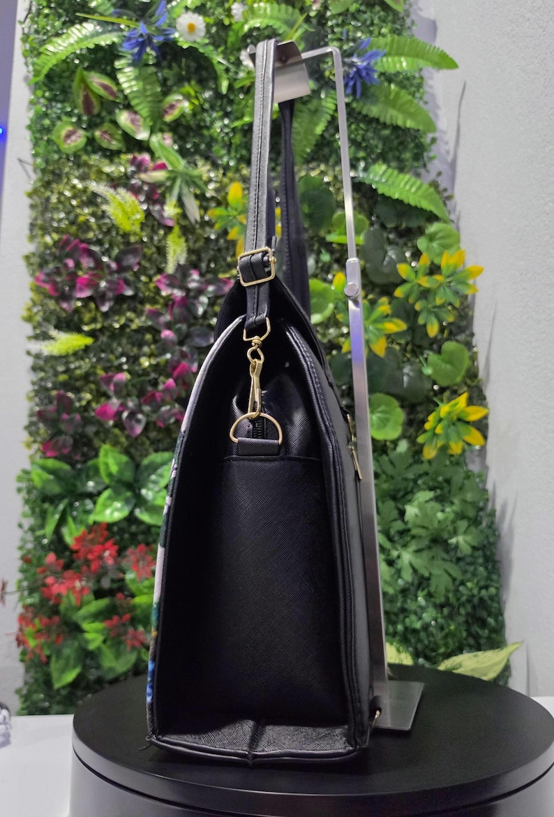 May include: A black handbag with a textured surface, featuring gold-toned metal hardware including a side D-ring, a swivel clasp, and an adjustable strap buckle. The bag is displayed in profile, showing its vertical orientation and structured side panel.