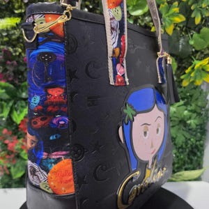 May include: A black tote bag featuring a Coraline character graphic and gold-toned lettering. The side panel displays a colorful pattern of keys, buttons, and characters. The bag includes adjustable straps with a multi-colored print and a black tassel.