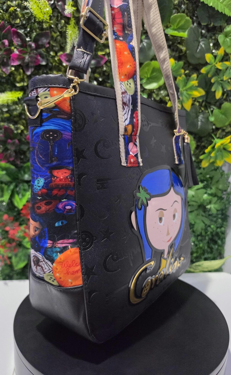 May include: A black tote bag featuring a Coraline character graphic and gold-toned lettering. The side panel displays a colorful pattern of keys, buttons, and characters. The bag includes adjustable straps with a multi-colored print and a black tassel.