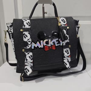 May include: A black satchel bag featuring a textured surface with a Mickey Mouse theme. The bag includes a top handle, a shoulder strap, and a front flap decorated with a holographic Mickey logo, a red bow, and two vertical white straps with black Mickey faces.