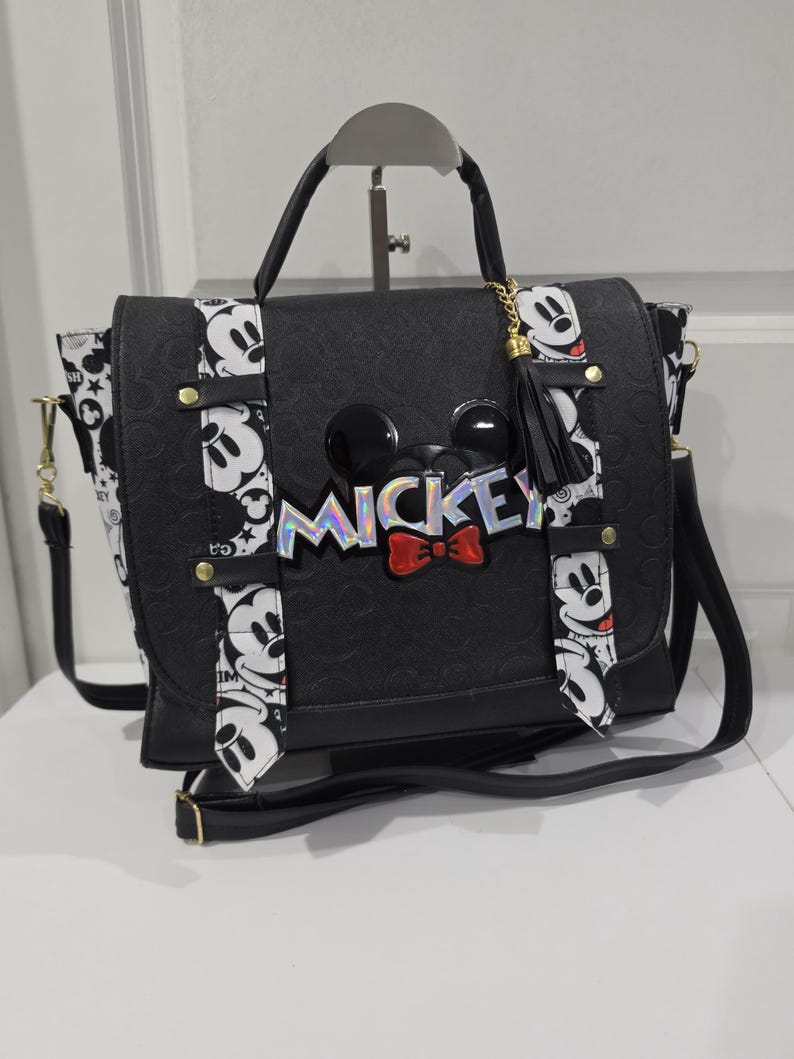 May include: A black satchel bag featuring a textured surface with a Mickey Mouse theme. The bag includes a top handle, a shoulder strap, and a front flap decorated with a holographic Mickey logo, a red bow, and two vertical white straps with black Mickey faces.