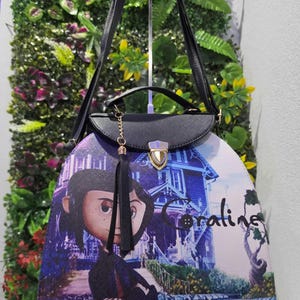 May include: A black handbag featuring a graphic print of the character Coraline standing in front of a blue house. The bag has a top handle, a shoulder strap, a gold-toned metal clasp closure, and a black tassel charm hanging from the front flap.