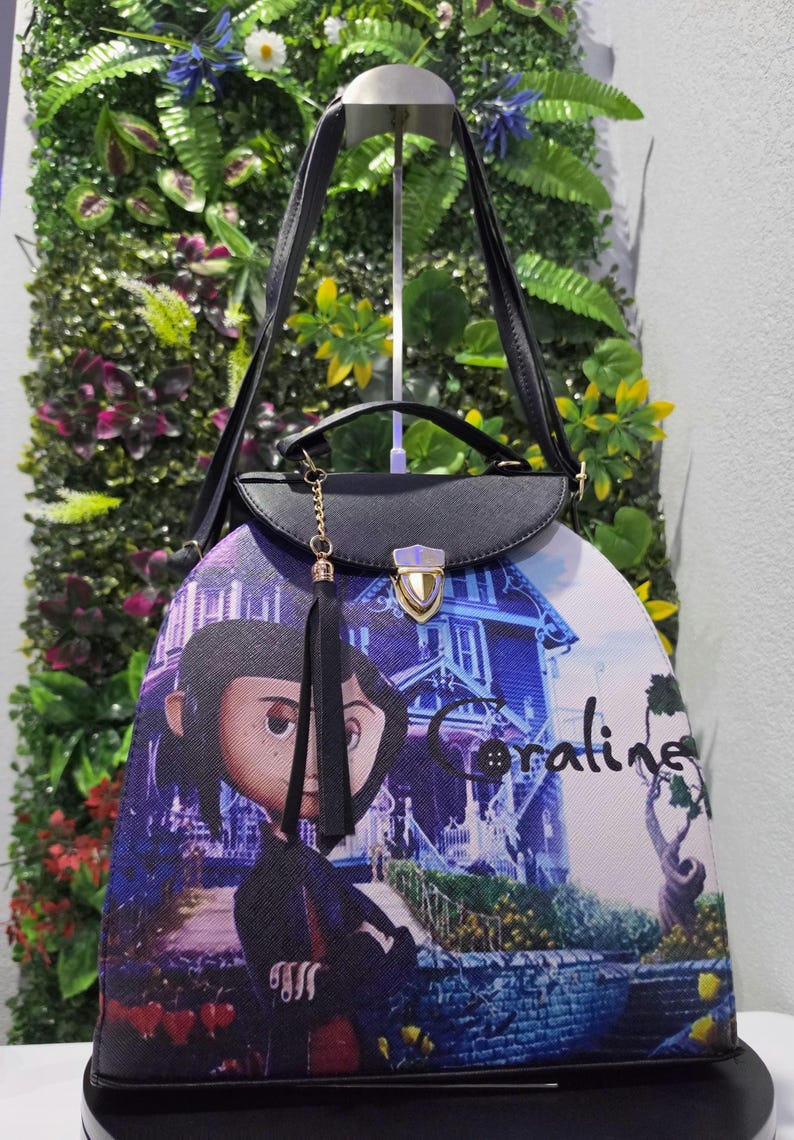 May include: A black handbag featuring a graphic print of the character Coraline standing in front of a blue house. The bag has a top handle, a shoulder strap, a gold-toned metal clasp closure, and a black tassel charm hanging from the front flap.