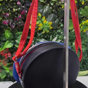 May include: A round crossbody bag featuring a black front panel, a blue middle section, and a white back panel. The bag includes bright red adjustable straps with white stitching, silver-toned metal hardware, and a zipper closure along the perimeter.