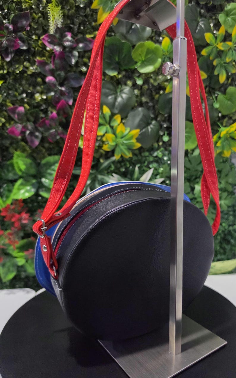 May include: A round crossbody bag featuring a black front panel, a blue middle section, and a white back panel. The bag includes bright red adjustable straps with white stitching, silver-toned metal hardware, and a zipper closure along the perimeter.