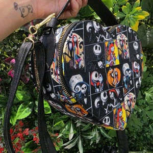 May include: A heart-shaped crossbody bag featuring a black fabric exterior with a repeating grid pattern of colorful illustrations depicting characters from The Nightmare Before Christmas, including Jack Skellington and Sally, with a black strap.