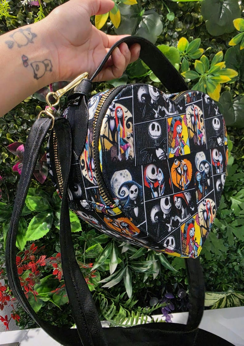 May include: A heart-shaped crossbody bag featuring a black fabric exterior with a repeating grid pattern of colorful illustrations depicting characters from The Nightmare Before Christmas, including Jack Skellington and Sally, with a black strap.