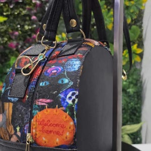 May include: A round black handbag featuring a colorful patterned fabric panel with various illustrations, including a bright orange circular graphic with the text Welcome home! in cursive script, black handles, and gold-toned metal hardware accents.