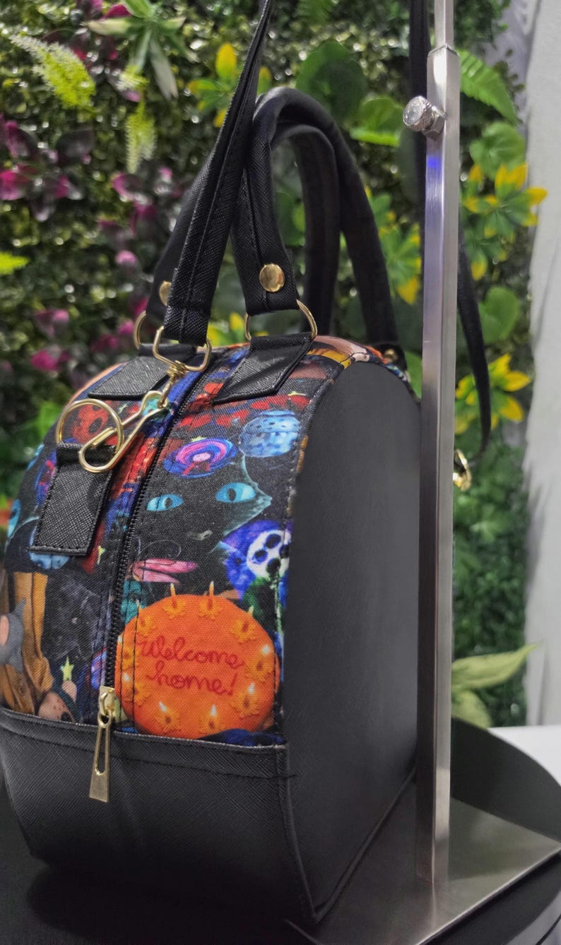 May include: A round black handbag featuring a colorful patterned fabric panel with various illustrations, including a bright orange circular graphic with the text Welcome home! in cursive script, black handles, and gold-toned metal hardware accents.