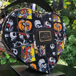 May include: A heart-shaped crossbody bag featuring a black, yellow, and white comic-style print of Jack Skellington and Sally from The Nightmare Before Christmas. The bag includes a black top handle, a shoulder strap, and a gold-tone metal plaque.