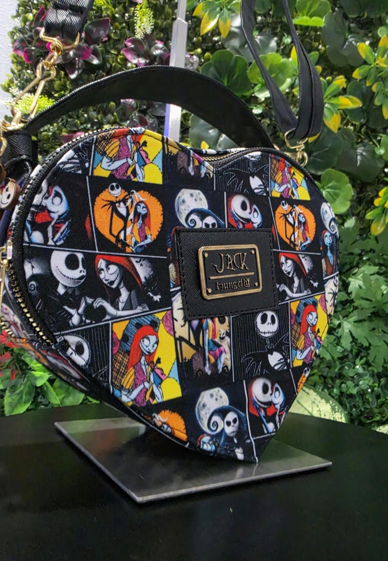 May include: A heart-shaped crossbody bag featuring a black, yellow, and white comic-style print of Jack Skellington and Sally from The Nightmare Before Christmas. The bag includes a black top handle, a shoulder strap, and a gold-tone metal plaque.