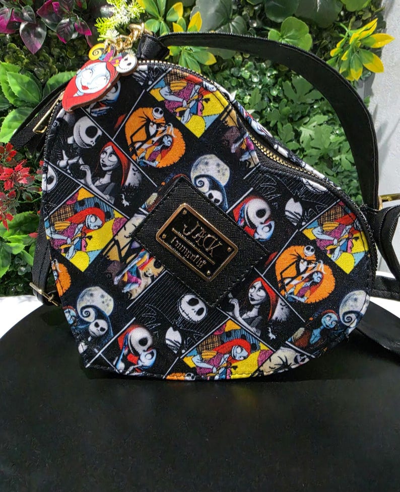 May include: A heart-shaped crossbody bag featuring a colorful all-over print of characters from The Nightmare Before Christmas. The bag has a black top handle, a gold-tone zipper, and a central black plaque with the text JACK and Loungefly.