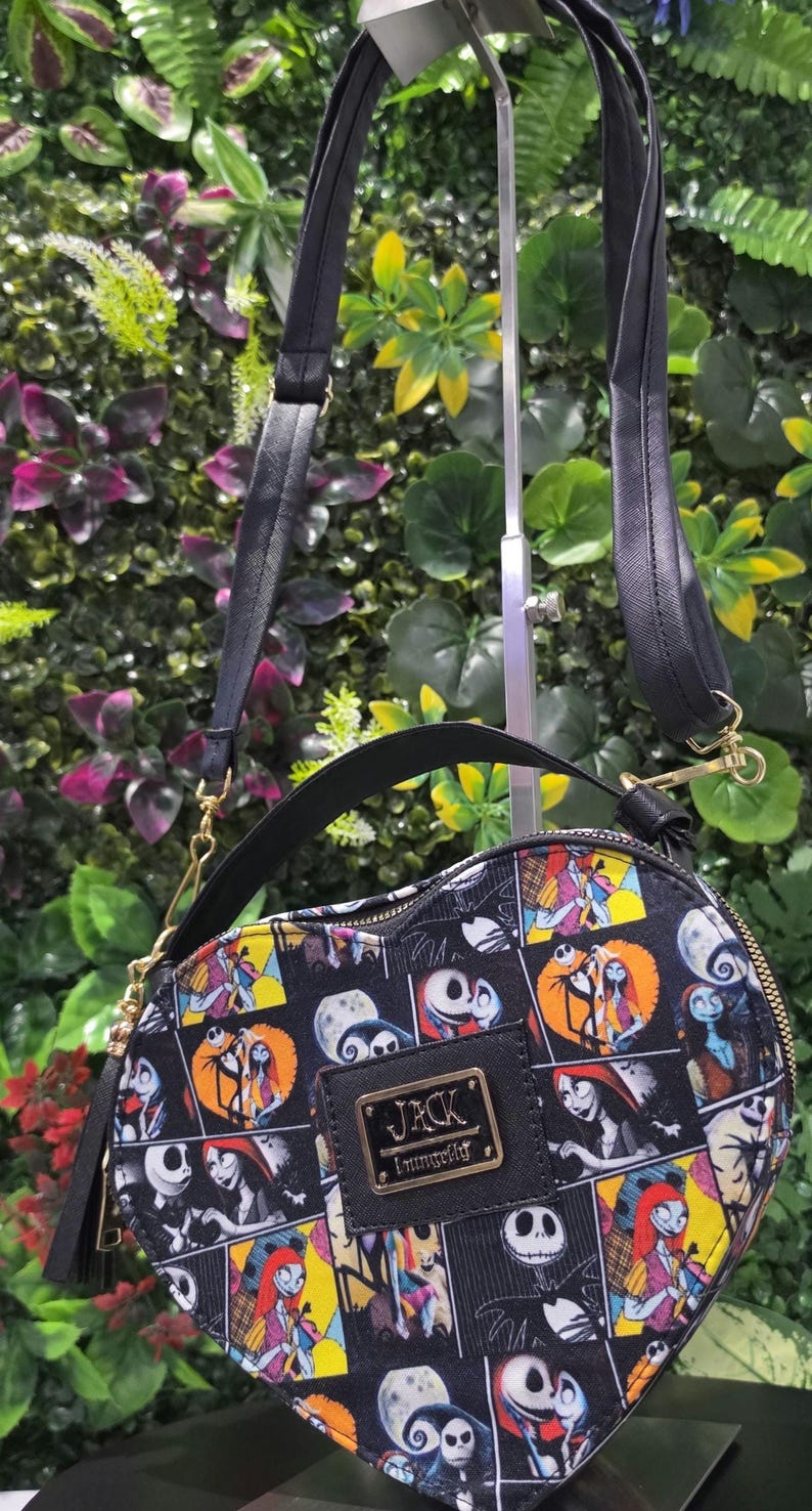 May include: A heart-shaped crossbody bag featuring a black and white comic-style print of characters from The Nightmare Before Christmas. The bag includes a black top handle, a detachable shoulder strap, and a gold-tone plaque reading JACK Loungefly.