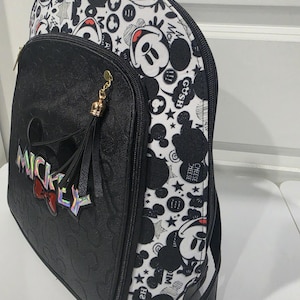May include: A black and white backpack featuring a repeating Mickey Mouse pattern on the sides and top. The front pocket is black with a debossed Mickey head design, a holographic MICKEY logo, a red bow accent, and a black tassel with gold hardware.