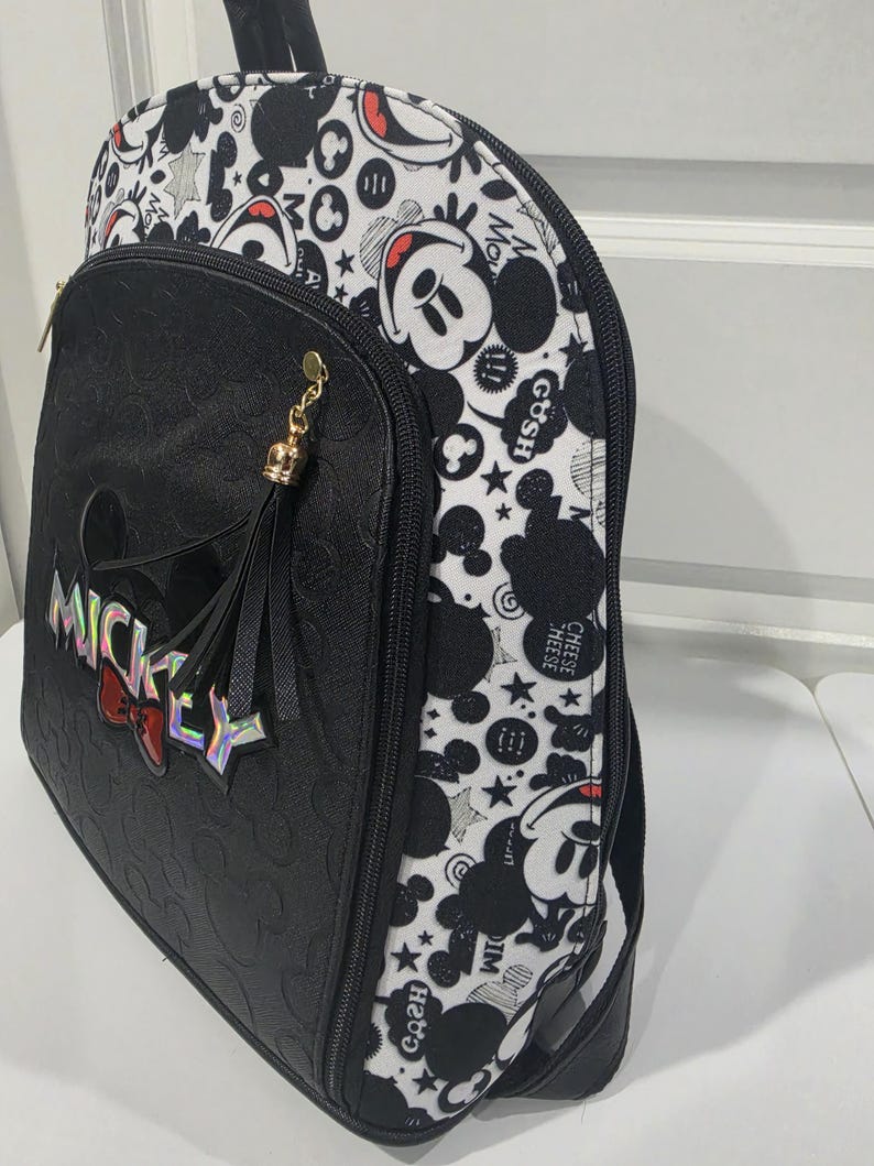 May include: A black and white backpack featuring a repeating Mickey Mouse pattern on the sides and top. The front pocket is black with a debossed Mickey head design, a holographic MICKEY logo, a red bow accent, and a black tassel with gold hardware.