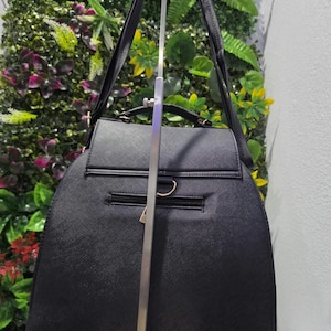 May include: A black structured backpack with a textured finish, featuring a top handle, adjustable shoulder straps, a horizontal zippered pocket on the back, and gold-toned metal hardware including D-rings at the base and a zipper pull.