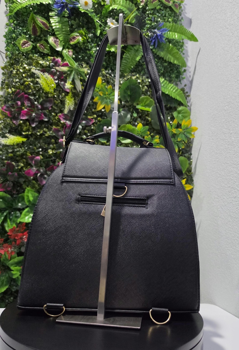May include: A black structured backpack with a textured finish, featuring a top handle, adjustable shoulder straps, a horizontal zippered pocket on the back, and gold-toned metal hardware including D-rings at the base and a zipper pull.