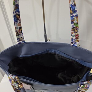 May include: A blue tote bag featuring a top zipper closure and a black interior lining. The bag has two shoulder straps decorated with a colorful cartoon character print. The exterior is a solid blue color with a visible zipper pull.