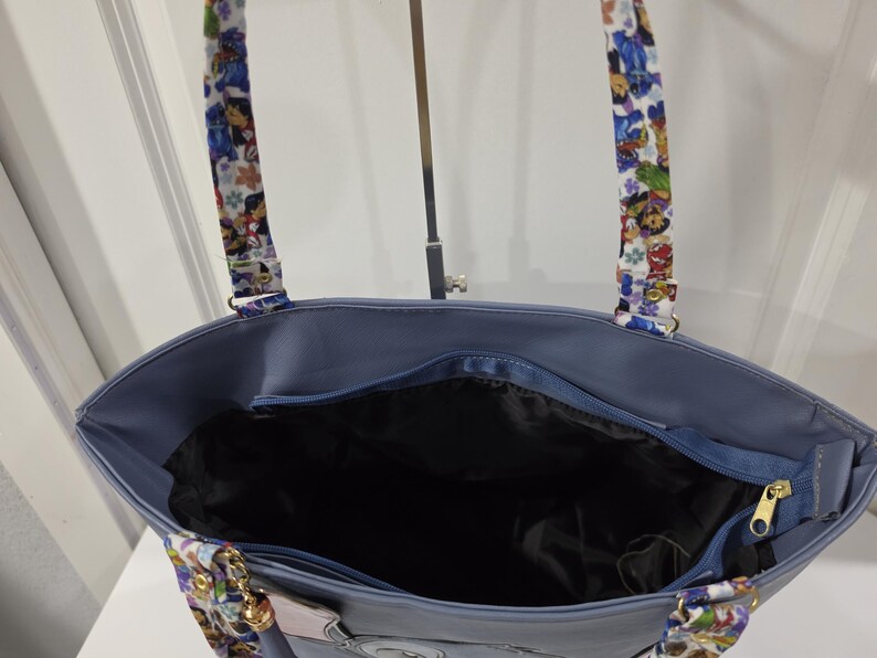 May include: A blue tote bag featuring a top zipper closure and a black interior lining. The bag has two shoulder straps decorated with a colorful cartoon character print. The exterior is a solid blue color with a visible zipper pull.