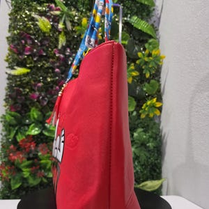 May include: A bright red tote bag featuring a cartoon character graphic on the side. The bag has dual handles with a blue, yellow, and white patterned design. A red tassel charm hangs from the base of one handle, and the bag has a textured finish.