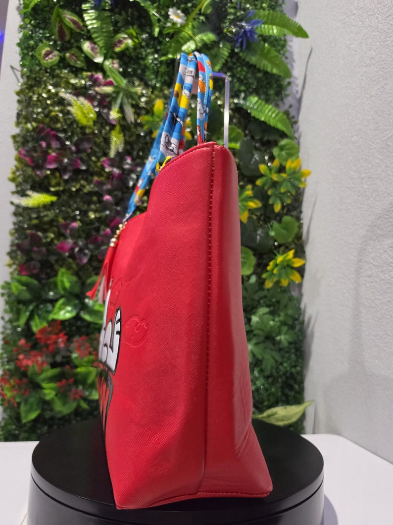 May include: A bright red tote bag featuring a cartoon character graphic on the side. The bag has dual handles with a blue, yellow, and white patterned design. A red tassel charm hangs from the base of one handle, and the bag has a textured finish.