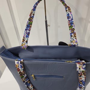 May include: A slate blue tote bag featuring a zippered top closure and a zippered exterior pocket with gold-toned hardware. The bag includes two shoulder straps decorated with a colorful repeating pattern of cartoon characters on a white background.
