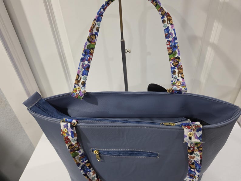 May include: A slate blue tote bag featuring a zippered top closure and a zippered exterior pocket with gold-toned hardware. The bag includes two shoulder straps decorated with a colorful repeating pattern of cartoon characters on a white background.